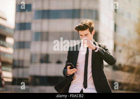 Junger Geschäftsmann Take away Kaffee trinken, während Smartphone Texte lesen Stockfoto