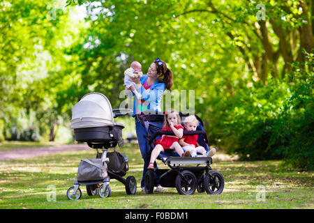 Junge Mutter in einem Park mit Kindern im Kinderwagen spazieren. Mutter und Kinder in einem Buggy Fuß im Wald. Frau drängen Doppel-Kinderwagen Stockfoto