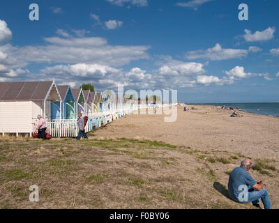 Menschen am Strand mit einer Reihe von bunten Strandhäuschen vor blauem Himmel. Mersea Island. Essex. England. VEREINIGTES KÖNIGREICH. Stockfoto