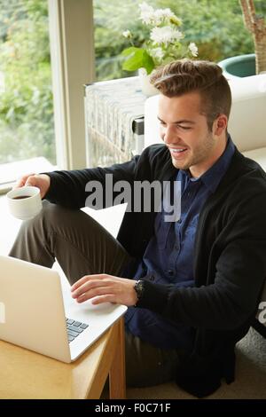 Porträt von lächelnder Mann auf Boden zu Hause sitzen mit Blick auf Laptop und Tasse Kaffee Stockfoto