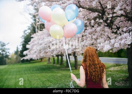 Rückansicht des junge Frau mit langen gewellten roten Haaren und Haufen Luftballons im Frühlingspark Stockfoto