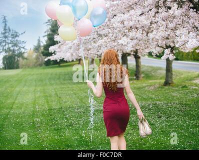 Rückansicht des junge Frau mit langen gewellten roten Haaren und Haufen Luftballons einen Spaziergang im Frühlingspark Stockfoto