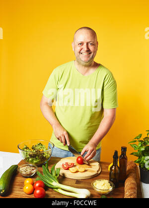 Frischer Salatzutaten auf den Tisch, von mittlerem Alter Mann geschnitten Tomaten - gelben Hintergrund Stockfoto