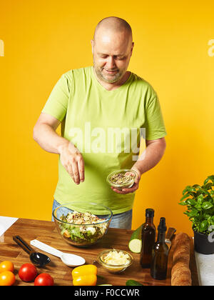 Frischer Salatzutaten auf den Tisch, von mittlerem Alter Mann streuen Samen - gelber Hintergrund Stockfoto