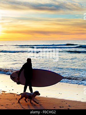 Silhouette eines Surfers mit einem Hund bei Sonnenuntergang am Strand. Portugal Stockfoto