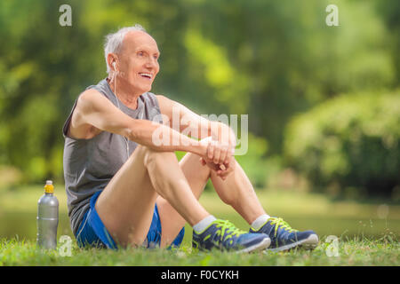 Sportliche Senior in Sportkleidung auf dem Rasen im Park sitzen und Musik über Kopfhörer hören Stockfoto