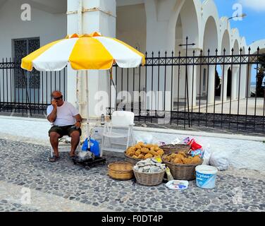 Ein Mann sitzt unter einem weißen und gelben Sonnenschirm, Schwämme und Bims-Steinen in Fira auf der Insel Santorin in Griechenland zu verkaufen Stockfoto