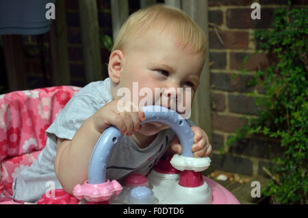 Ein Zahnen zehn Monate altes Baby sitzen in ihrer Gehhilfe beißen auf die Kunststoff-Griff, die Schmerzen und Beschwerden des Zahnes zu lindern. Stockfoto