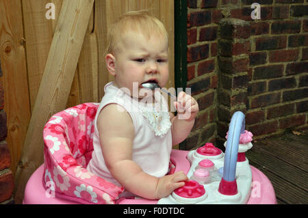 Ein Zahnen zehn Monate altes Baby sitzen in ihrer Gehhilfe beißen auf einem Löffel, die Schmerzen und Beschwerden von Zahnschmerzen zu lindern. Stockfoto