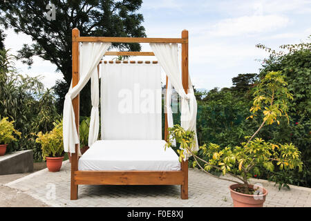 Outdoor-Holzbett mit weißer Bettwäsche und Vorhängen auf einen Backstein Innenhof in einem tropischen Resort. Stockfoto