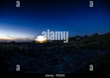 Wie Dämmerung über eine Bergkette absetzt, ist ein Zelt gegen einen Sternenhimmel mit Lichtern aus dem Tal unten noch glühende beleuchtet Stockfoto