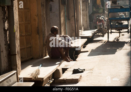 Amritsar, Punjab, Indien. Mann sitzt barfuß auf den Schritt seines Hauses mit einem kurzen geschwungenen Besen und seine Schuhe auf der Treppe. Stockfoto