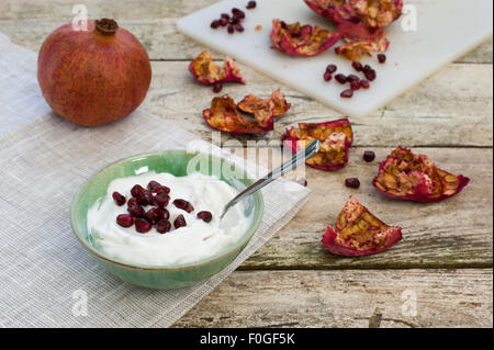 Schüssel mit Joghurt, garniert mit Granatapfelkernen auf rustikalen Tisch mit ganzen Granatapfel, Schale und Samen im Hintergrund Stockfoto