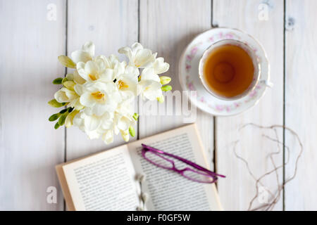 Nahaufnahme des weißen Freesien auf blauen Tischset mit unscharfen Vintage Teetasse und Untertasse im Hintergrund, auf rustikalen Holztisch Stockfoto