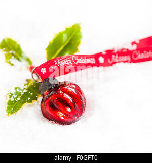Runde Rote Christbaumkugel mit Holly und Frohe Weihnachten Ribbon auf Schnee Stockfoto