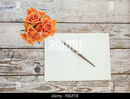 drei orange Rosen auf rustikalen Holztisch mit offenen Notizbuch und Stift Stockfoto