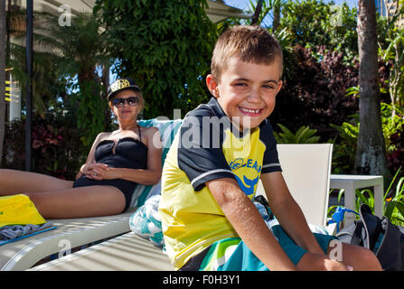 Junge mit mit breiten Grinsen am Pool in einem Ferienort. Seine gut aussehende Mutter in ihren Vierzigern blickt auf Entspannung. Stockfoto