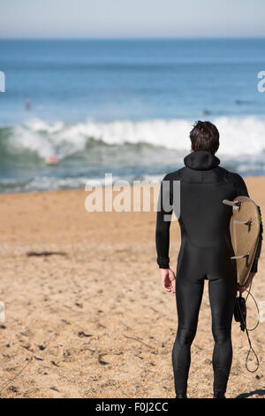 Junge attraktive Surfer Mann stehend an einem Strand und tragen seine Surf-Board beim Blick auf den Atlantischen Ozean während ein sunn Stockfoto