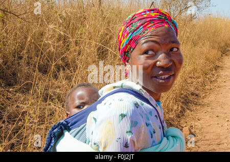 Eine Mutter trägt ihren kleinen Sohn auf dem Rücken, wie sie durch die Feldwege von ländlichen Swasiland Wanderungen. Stockfoto