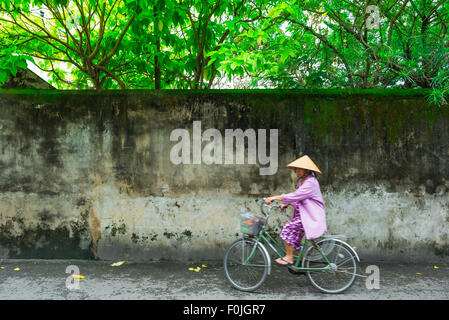 Vietnam-Frau radelt, Blick auf eine ältere Vietnamesin, die einen konischen Hut trägt und in rosa gekleidet ist, die auf einer Straße in Hoi an, Zentralvietnam, fährt. Stockfoto