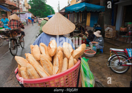 Vietnam Bread, Rückansicht einer Händlerin, die einen Korb mit französischem Brot - Banh mi - auf den Lebensmittelmarkt in Hoi an, Zentralvietnam liefert. Stockfoto
