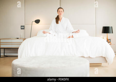 Gelassene Frau im Bademantel meditieren im Lotussitz auf Bett Stockfoto