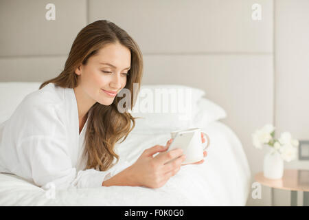 Lächelnde Frau im Bademantel, trinken Kaffee und SMS auf dem Handy auf Bett Stockfoto