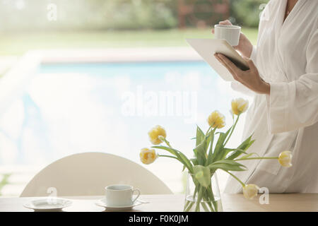 Frau im Bademantel, Kaffee trinken und mit digital-Tablette am Pool Stockfoto