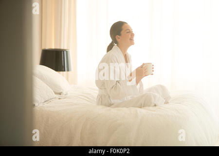 Lächelnde Frau in Bademantel Kaffeetrinken im Schneidersitz auf Bett Stockfoto