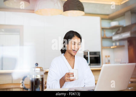 Lachende Frau im Bademantel, Kaffee trinken und mit laptop Stockfoto