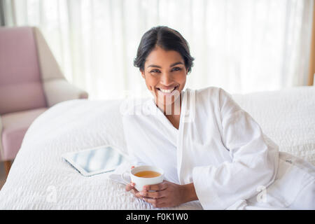 Lächelnde Frau Porträt im Bademantel Teetrinken auf Bett Stockfoto