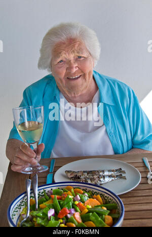 LANGLEBIGKEIT Happy ältere Dame 90s im Urlaub genießen gesunde Mahlzeit unter freiem Himmel mit frischem gegrilltem Fisch, gemischtem Bohnensalat und einem Glas Weißwein Stockfoto