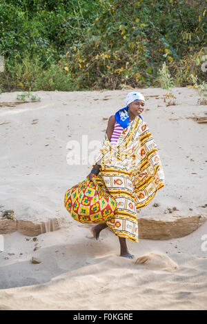 Lokale junge afrikanische Frau im bunten lokalen Kleid zu Fuß am sandigen Ufer auf der angolanischen Seite der Grenze zu Namibia, tragen ein Tuch-bundle Stockfoto