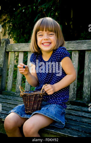 Kleines Mädchen sitzt auf einer Gartenbank mit Wickerbasket Blaubeeren grinsend Stockfoto