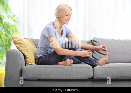 Junge Frau sitzt Polieren ihre Fußnägel und lächelnd auf eine graue Couch zu Hause Stockfoto