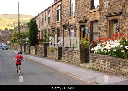 Ein Junge spielt auf einem Roller durch Reihenhäuser Kinder unterwegs in Hayfield Dorf, Peak District, Derbyshire, England UK Nord Stockfoto