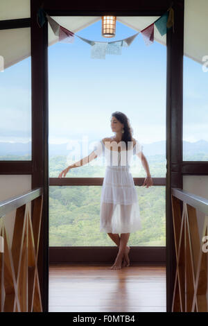 Junge Frau in einem weißen Kleid in einer meditativen stehend Pose und Übung Achtsamkeit in einem Türrahmen mit blau Himmel und Wald hinter ihr Stockfoto