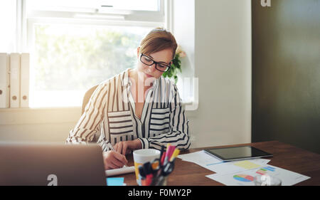 Hübsche junge Geschäftsfrau schreiben einige Notizen auf ihren Schreibtisch in the Office gegen the Glasfenster. Stockfoto