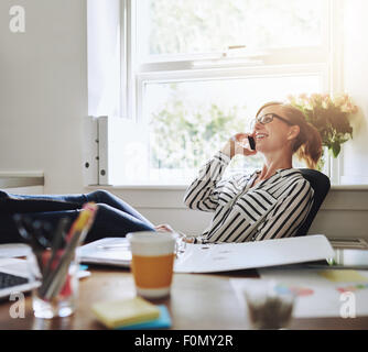 Glückliche junge Geschäftsfrau im Gespräch mit jemand auf ihr Handy während entspannende Inside the Office mit Beinen auf ihrem Schreibtisch. Stockfoto