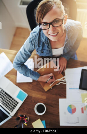 Junge Frau lächelt in die Kamera von hohen Winkel Ansicht während machen einige Notizen auf Papier Geschenktüte am Holztisch hautnah. Stockfoto