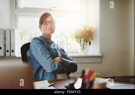 Selbstbewussten Jungen Büro Frau sitzt an ihrem Schreibtisch mit Arme über ihren Bauch, in die Kamera lächeln gegen das Glas Wi Stockfoto