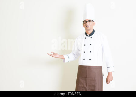 Porträt der schönen indischen männlichen Koch in einheitliche begrüßen und lächelnd, stehend auf einfarbigen Hintergrund mit Schatten, Textfreiraum auf Stockfoto
