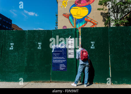 New York, NY - 14August 2015 - A Passant guckt durch den Zaun zu brasilianischen Twin Straßenkünstler Os Gemeos sehen erstellen eine 6 Geschichte Wandbild im Stadtteil East Village von Manhattan Stockfoto