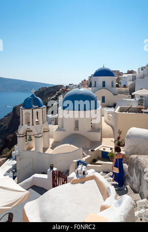 Junge Frau geht pass blaue Kuppel-Kirche des Agios Spiridonas und Kirche von Anastasis (Auferstehung) in Oia, Santorini, Griechenland Stockfoto