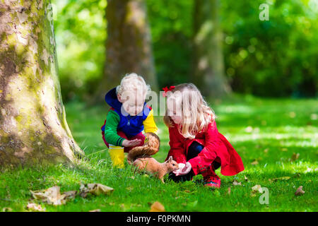 Kinder füttern Eichhörnchen im Herbst Park. Kleine Jungen und Mädchen im roten Mantel und Regen Stiefel sehen Sie wilde Tiere im Herbst Wald Stockfoto