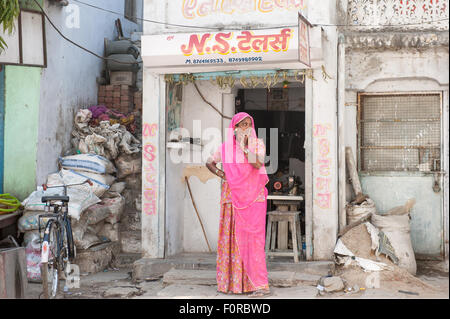 Rajasthan, Indien. Zwischen Jodhpur und Jaipur. Frau stehend außerhalb ihrer kleinen Näherei mit ihrer Nähmaschine in traditionellen Rajasthan rosa Sareelooking entlang der Straße mit ihren Fingern über den Mund. Stockfoto