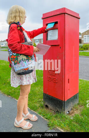 Ältere Frau mailing einen Brief in einen roten rechteckigen Briefkasten in England, Großbritannien, Posten Briefe. Buchen einen Brief. Stockfoto