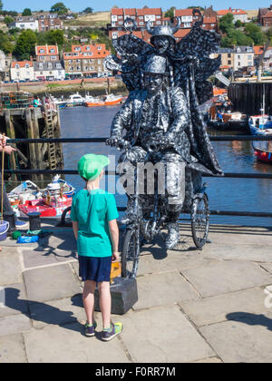 Kleiner Junge beobachtet einen Mann posiert als lebende Statue auf einem Dreirad und mit einem Teufel auf dem Rücken in Whitby North Yorkshire England Stockfoto