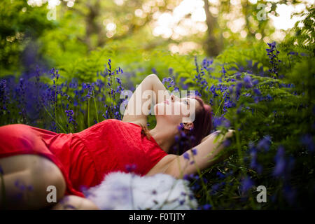 Eine junge Frau Teenager-Mädchen trägt ein rotes Kleid allein in einem Waldgebiet liegend in einem Feld von Glockenblumen auf Frühjahr Abend UK Stockfoto