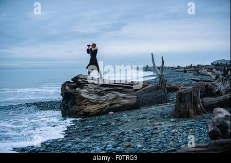 Frau, die durch ein Fernglas aus großen Treibholz Baumstumpf am Strand Stockfoto